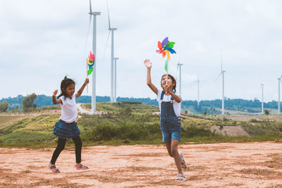 Full length of sisters playing with pinwheel toys on land with windmills in background