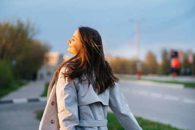 Beautiful smiling girl with long hair in a grey trench coat outdoors on the street spring