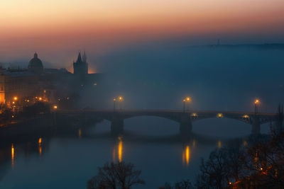 Illuminated bridge over river against buildings during sunset