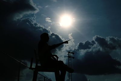 Low angle view of silhouette man pointing while sitting on chair against cloudy sky on sunny day