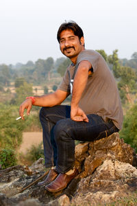 Full length portrait of man smoking while sitting on rock