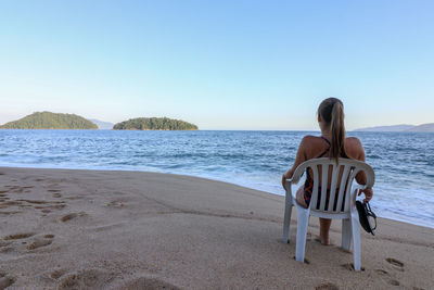 Rear view of woman sitting on chair at sandy beach against clear sky