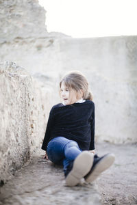 Playful girl looking away while sitting by retaining wall
