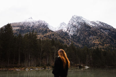 Back view of unrecognizable female tourist enjoying amazing landscape of calm lake and majestic mountains with forested slopes and snowy peaks in cloudy autumn day