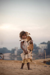 Portrait of girl carrying dog while walking on sand against sky