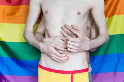 Midsection of shirtless gay couple embracing against rainbow flag