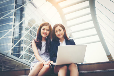 Portrait of happy young friends with laptop sitting steps in city