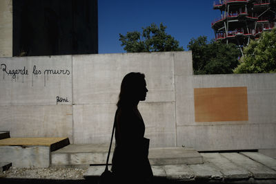 Side view of silhouette woman standing on sidewalk with graffiti on wall