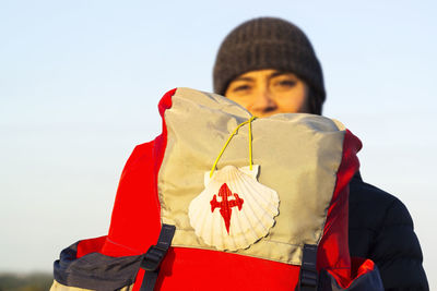Portrait of young woman holding backpack while standing against sky