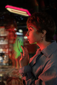 Lonely millennial girl drawing heart with finger on shop window, standing in neon light at night