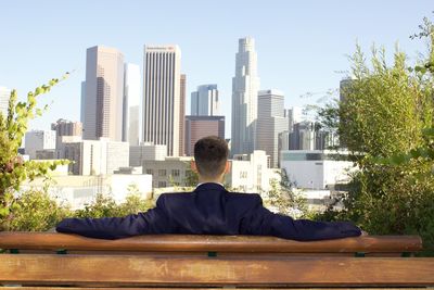 Rear view of businessman sitting on bench against towers in sunny day