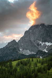 Scenic view of a rocky mountain peak with a hut surrounded by trees at sunset in a mountain