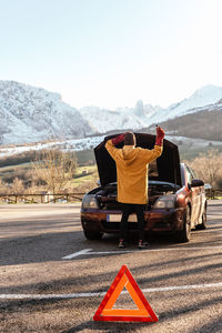 Back view full length person in outerwear opening car hood parked near warning triangle road sign in sunny winter highlands person