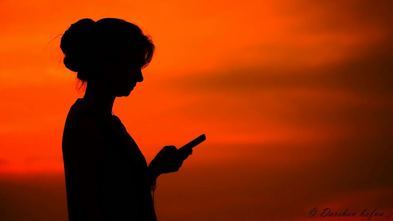Side view of silhouette woman using phone against orange sky at dusk