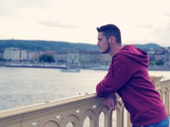 Side view of thoughtful young man looking away while standing on bridge in city