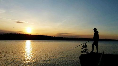 Silhouette man fishing by lake against sky during sunset