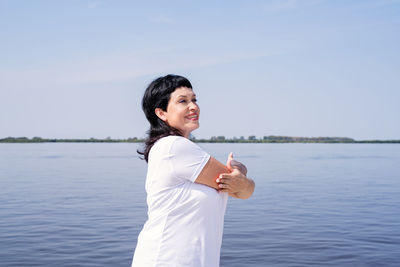 Smiling young woman standing by lake against sky