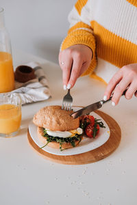 Unrecognizable woman eating breakfast sandwich with egg and arugula and cherry tomatoes on the tabl