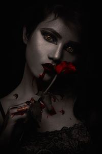 Close-up portrait of spooky young woman holding red rose against black background