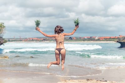 Rear view of woman in bikini holding pineapples while jumping against sea at beach