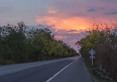 Road by trees against sky during sunset