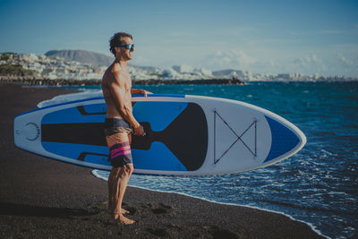 A young sportsman man practices paddle surfing on the beach under a blue sky