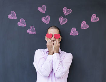 Man with heart shape paper on eyes standing against blackboard