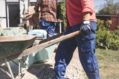 Side view of senior woman pushing wheelbarrow at yard while man standing in background