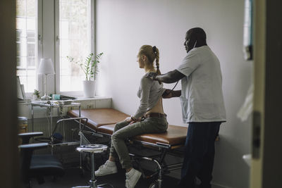 Side view of mature male doctor examining female patient sitting on bed in clinic