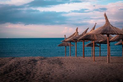 Thatched roofs on beach against cloudy sky during sunset