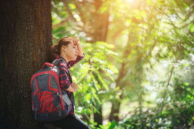 Tired female hiker carrying backpack while leaning on tree trunk in forest