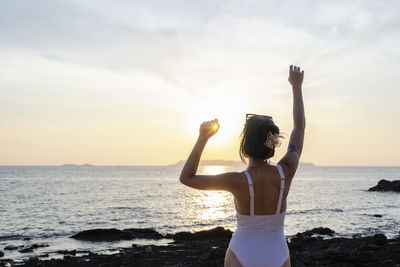 Rear view of woman with arms raised standing at beach against sky during sunset