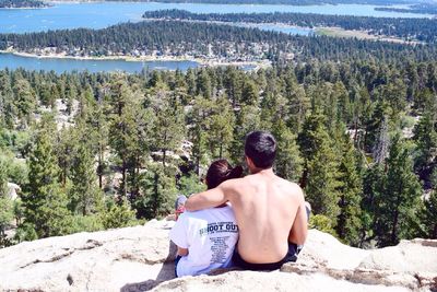 Rear view of couple sitting on rock against trees during sunny day