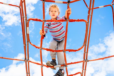 Low angle view of boy on red ropes against blue sky