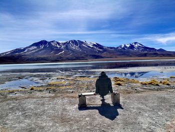 Rear view of woman sitting at lakeshore against snowcapped mountains