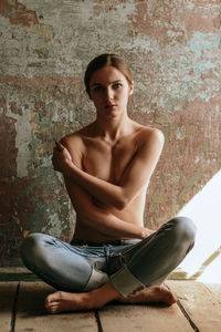 Portrait of shirtless young woman sitting on floor against wall