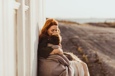 Mother kissing while sitting with son by built structure