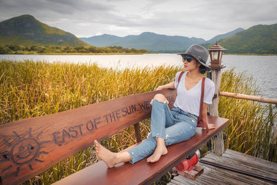 Young woman sitting on bench by lake against mountains