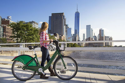 Woman with bicycle walking on bridge looking at river against city