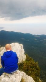 Rear view of man sitting on mountain by sea against sky