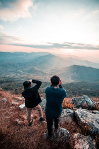 Rear view of friends photographing through camera while standing on mountain against sky during sunset
