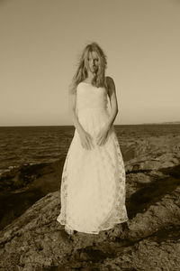 Sad young woman wearing white dress standing at beach against sky