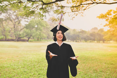 Young woman in graduation gown playing with certificate while standing on grassy field at park