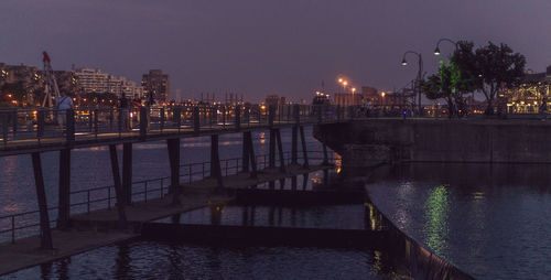 Illuminated bridge over river by buildings against sky at night