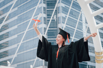 Happy young woman wearing graduation gown while standing against office building