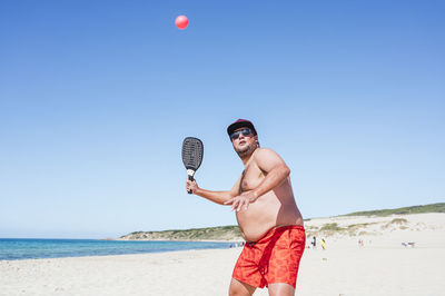 Overweight man playing paddle ball at beach during sunny day