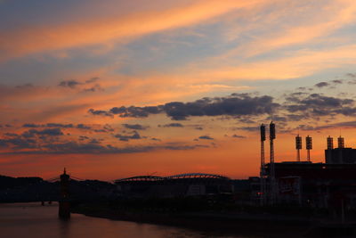 Silhouette factory by buildings against sky during sunset