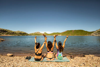 Back view of group of female friends in swimwear having fun and raising arms while enjoying summer journey on shore of mountain lake in pyrenees