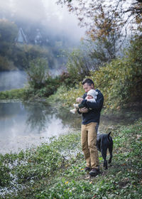 A man is holding a baby near a river and a dog