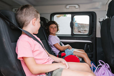 Brother and sister in casual clothes looking at each other while sitting on back seat of vehicle during road trip